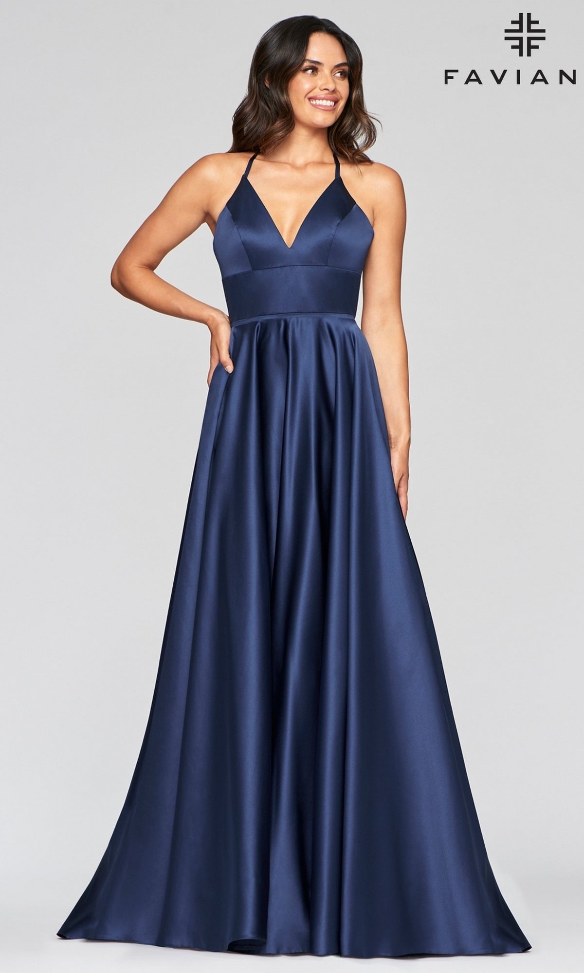 A woman models the Corset-Back A-Line Long Satin Prom Dress by Faviana in navy blue satin with a deep V-neck and thin straps, standing before a plain light background. The Faviana logo appears in the upper right corner.